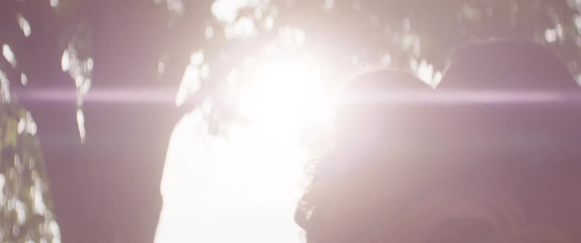 Sun glares onto a mother turning in circles under the trees while holding her daughter in her arms
