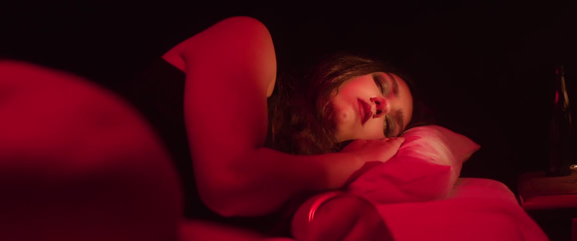 A young woman sleeps on a bed in a room illuminated by deep red light as another light briefly reveals her face