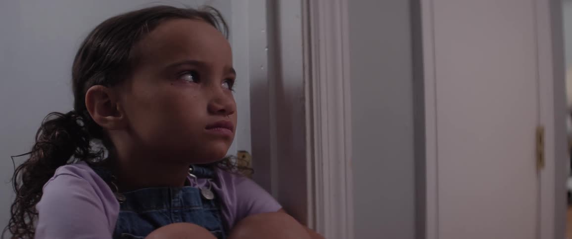 A little girl sits in an empty room against a wall looking fearful and upset
