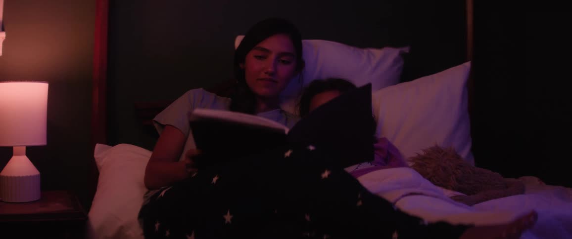 An older sister reads a book to her younger sister in bed under warm ambient light