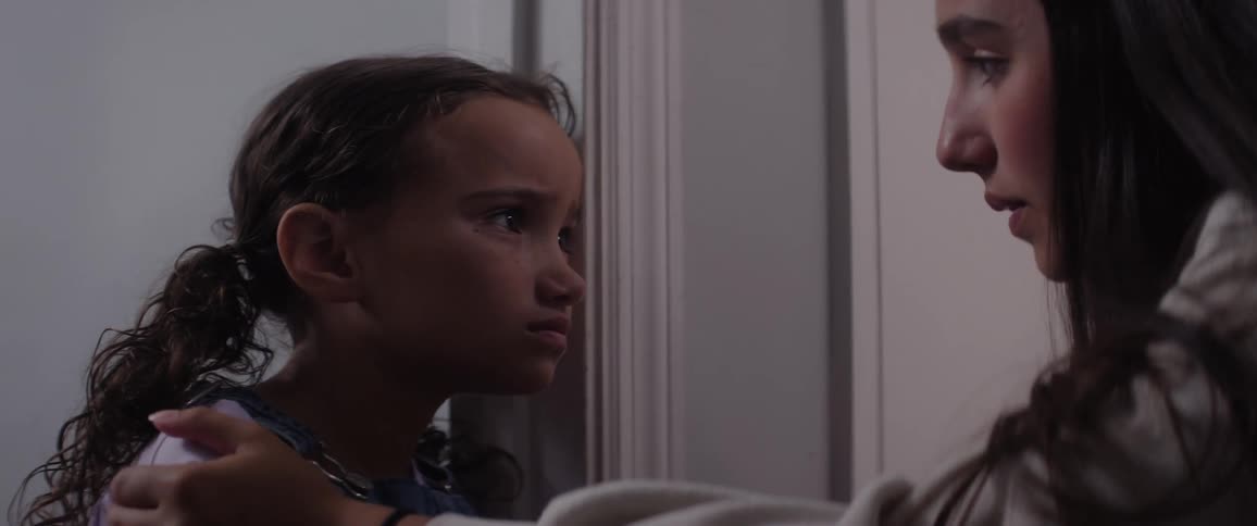 A teenage girl walks over to a young child standing sadly beside a door in their house and gently rubs her shoulder to cheer her up