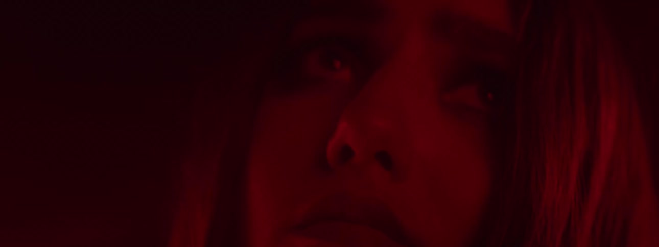A woman looks upwards with a serious expression her face illuminated by strong red light in a dark environment