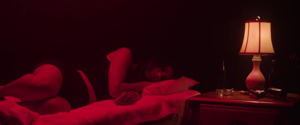 A brief light reveals the face of a young woman sleeping on a bed in a room illuminated by dark red fluorescent light with a lampshade on the side