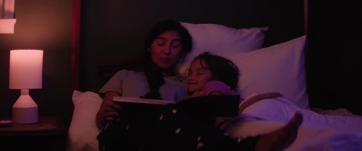 A young woman reads a book to a child snuggled beside her in bed illuminated by a warm bedside lamp