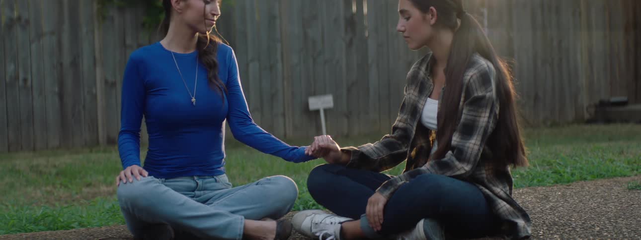 Two women with long dark hair are seated on green grass facing each other with a wooden fence in the background