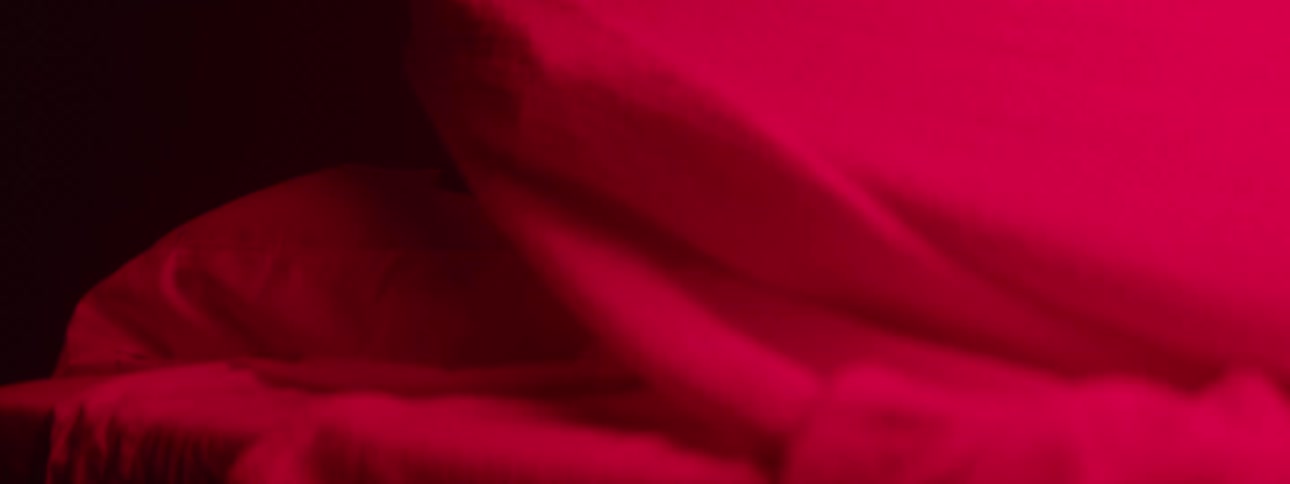 A young woman in her bedroom at night illuminated by red ambient light and a lamp behind her uses a blanket to cover her face as she tries to fall asleep