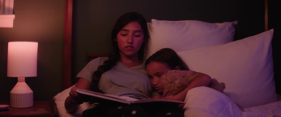 Mother sits on a bed reading a book aloud as her daughter listens clutching a stuffed animal