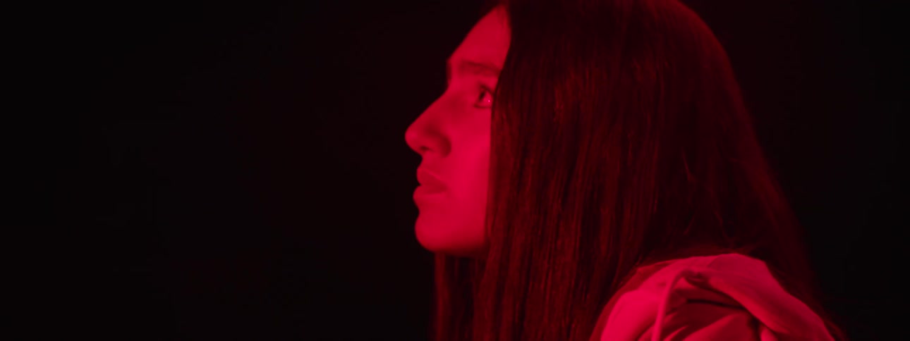 A young woman sits in a dark room looking nervous as red light illuminates the atmosphere