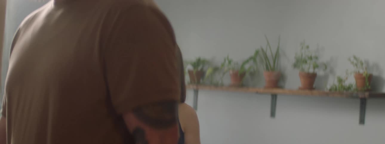A woman with a sad expression sits in a room with clasped hands facing a tattooed man as they talk