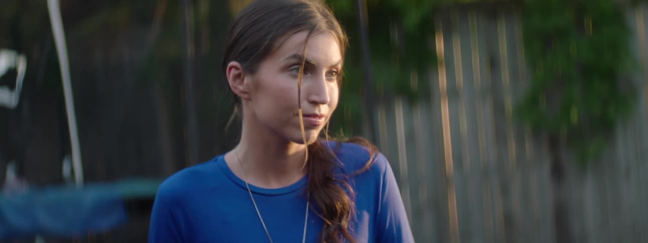 A woman wearing a blue shirt and necklace sits in front of a weathered wooden fence talking to someone