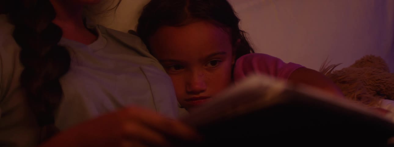 An older sister reads a book to her younger sister in bed under warm ambient light