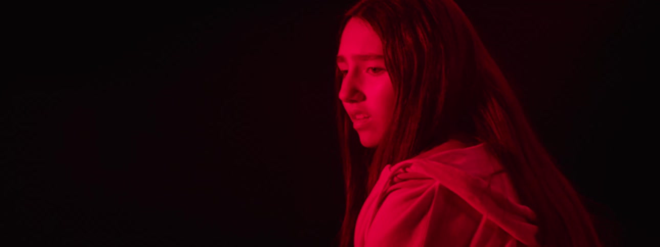 A teenage woman with long dark hair is illuminated by a strong red light looking over her right shoulder with a wide eyed expression in a dark environment