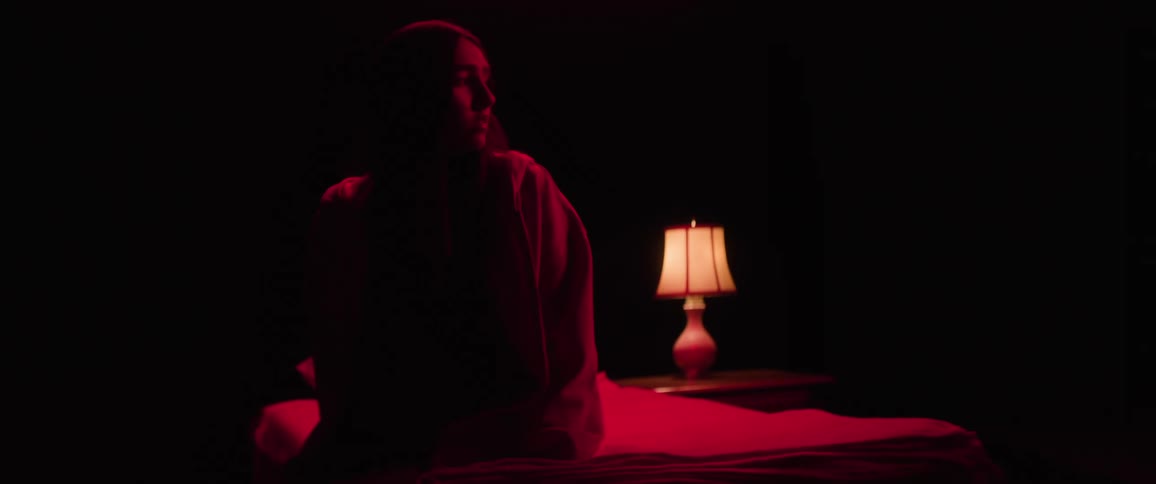 A young woman sits on the side of a bed in a dark room illuminated by a red fluorescent light with a lamp beside her