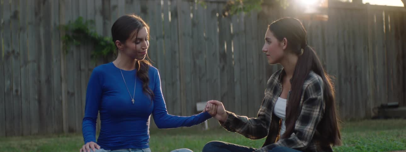 Two young women sit in a backyard holding hands in a quiet moment