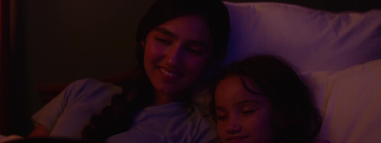 Female teen lies on the bed to read a book to her younger sister at night before bedtime