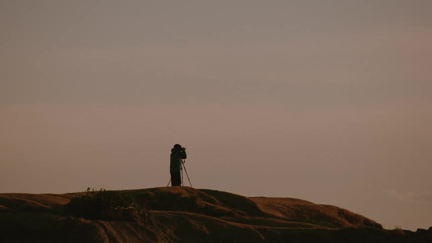 Photographer stands on a rocky hilltop with a tripodmounted camera capturing the landscape under a vivid orange sky