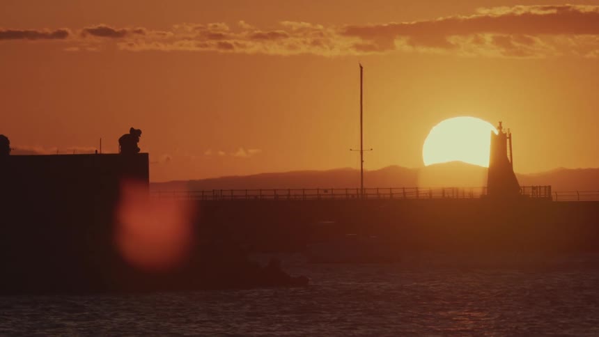 A person stands silhouetted on a pier at sunset with an orange sky and a lighthouse in the background