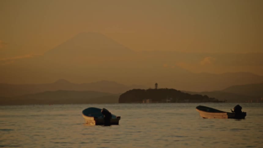 Two small boats float on calm water with a large mountain and a smaller island with a tower visible in the distant background under an orange sky