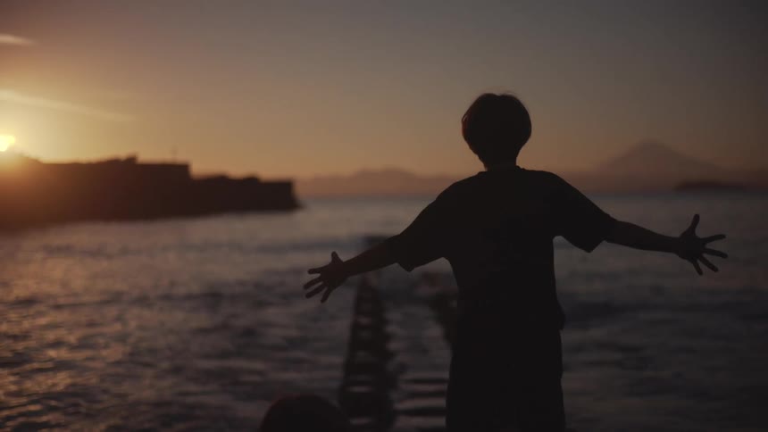 A child dances along a riverbank at sunset with the orange setting sun visible