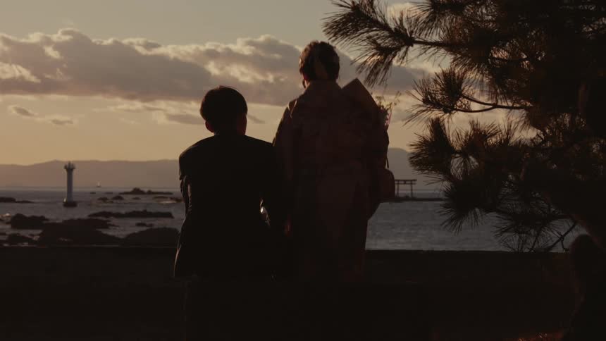A man and a woman wearing a traditional japanese kimono sit silhouetted by the ocean at sunset with a lighthouse and low clouds visible in the distance