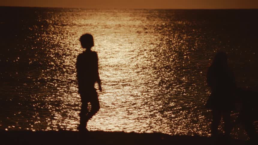 Three kids silhouettes walk along the beach with sunlight glistening on the ocean