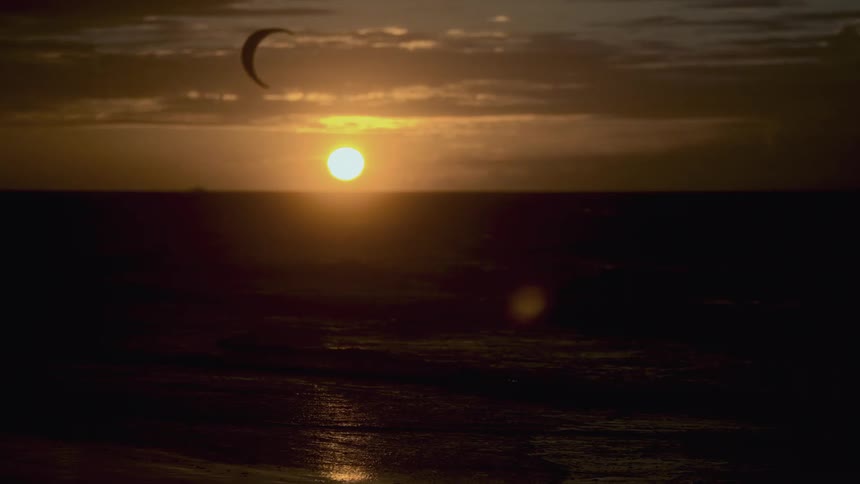 A large bright orange sun sets over a calm ocean with a crescent shaped kiteboarding parachute visible in the sky above