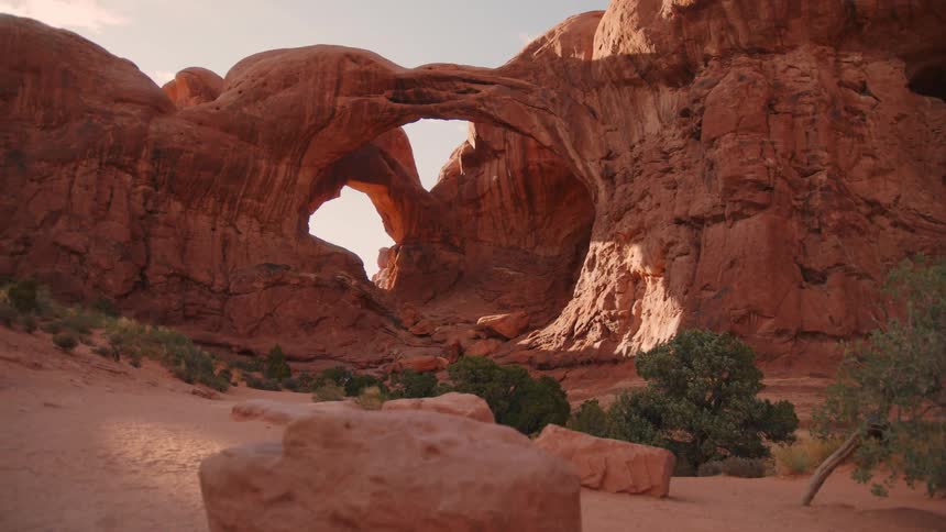 A natural sandstone arch rises from rocky terrain surrounded by desert shrubs under a partly cloudy sky