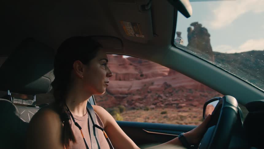 A woman with long brown hair drives a car looking out the window at a desert landscape with rock formations under a bright sky