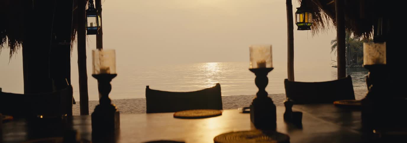 A sunset lit seaside dining area overlooks the calm ocean from a shaded terrace