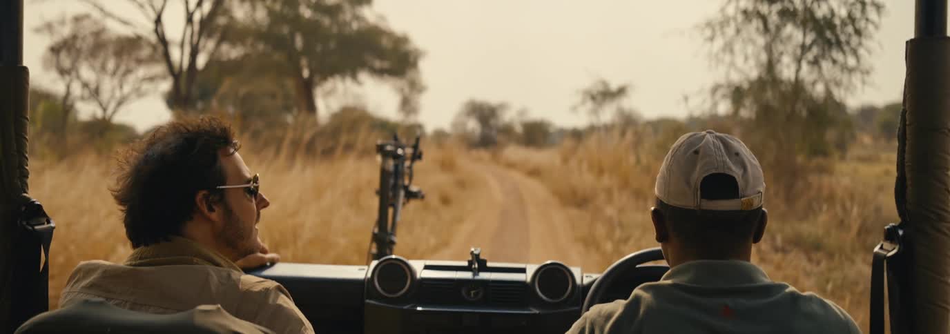 Two men drive in a car down a dirt path through a grassy field