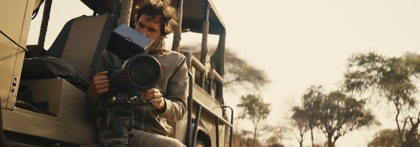 A person holding a large camera sits on the back of an open vehicle parked near tall trees under a clear sky
