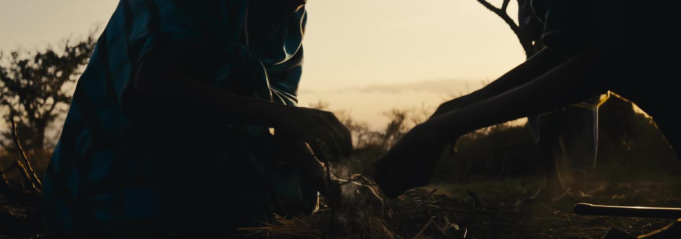 Two people in tribalstyle clothing prepare a small fire near a tree line at sunset