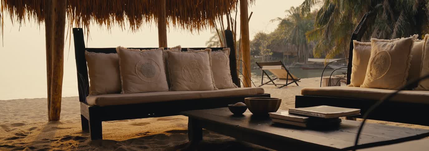 A shaded outdoor seating area with a wooden sofa and table holding bowls and books overlooks a sandy beach and calm water with distant palm trees and lounge chairs