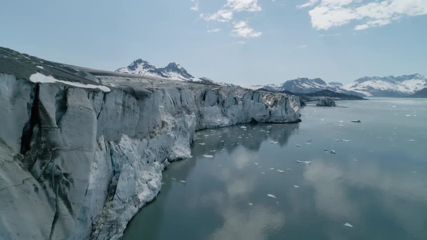 A dramatic aerial view of a glacier cliffside meeting a calm icy coastal bay under a bright sky