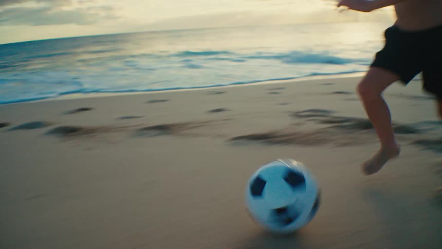 Boy running on the shoreline kickng a soccer ball at sunset