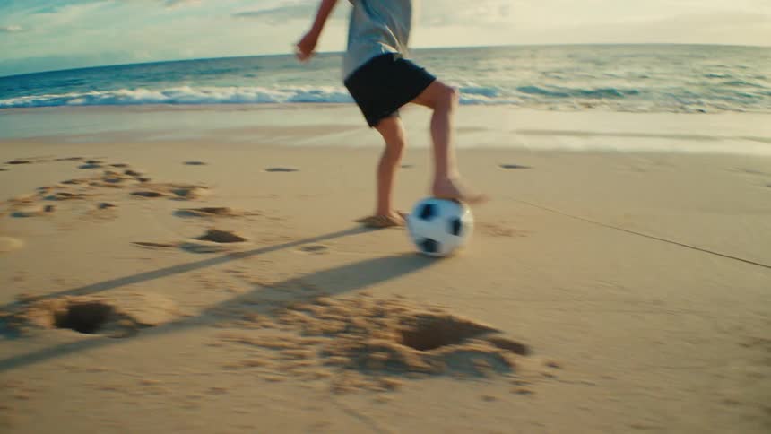 A young boy plays barefoot with a soccer ball on a sandy beach near the ocean