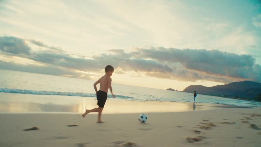Boy running and kicking a soccer ball on the beach as waves roll in to shore