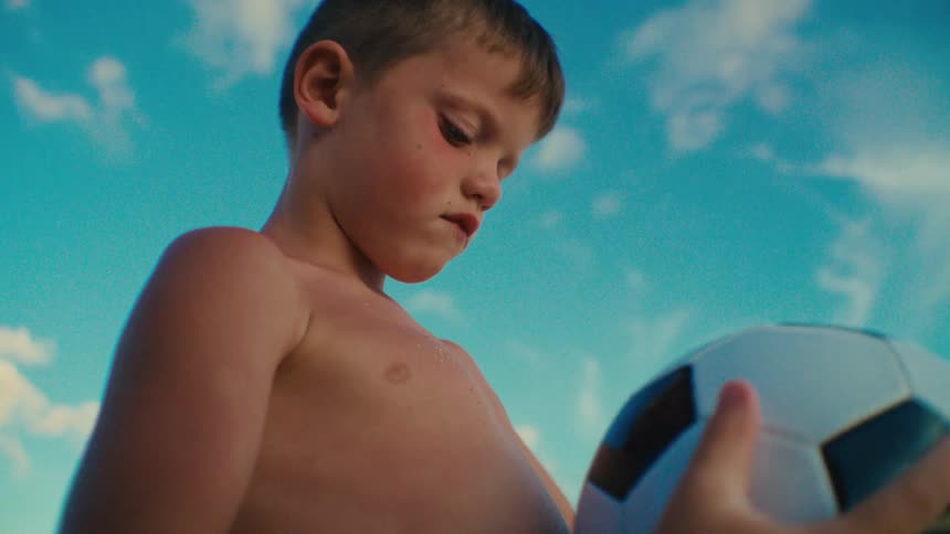 A shirtless child stands outdoors holding a soccer ball