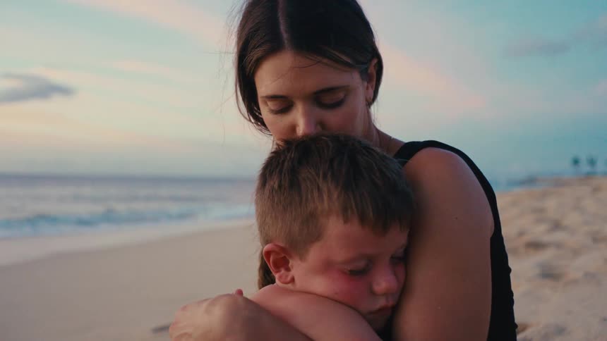 Mother hugs and kisses her sun while sitting on the sand at the beach during daytime