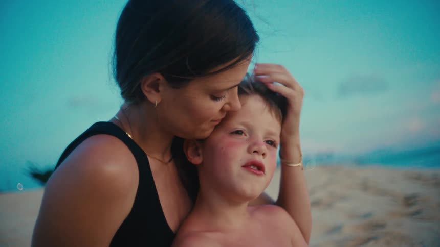 A mother kisses her young son on the beach at sunset