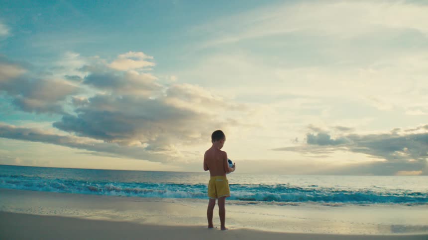 Back of young boy holding soccer ball standing on shoreline at the beach at sunset