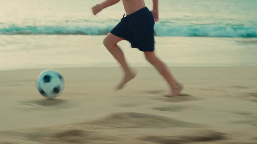 A young boy runs while kicking a soccer ball on the beach during daytime