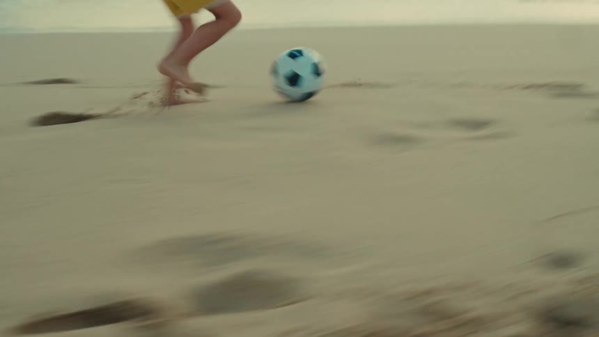 A young boy plays soccer alone on a beach at sunset