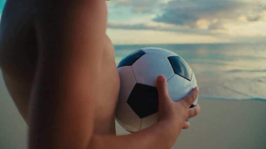 Close up of a boy inspecting a soccer ball while standing at the beach at sunset