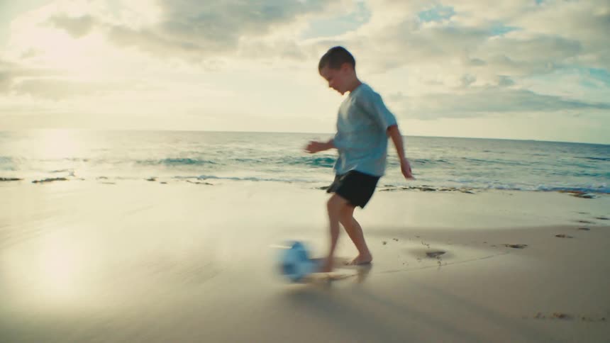 The boy is kicking a soccer ball on the beach