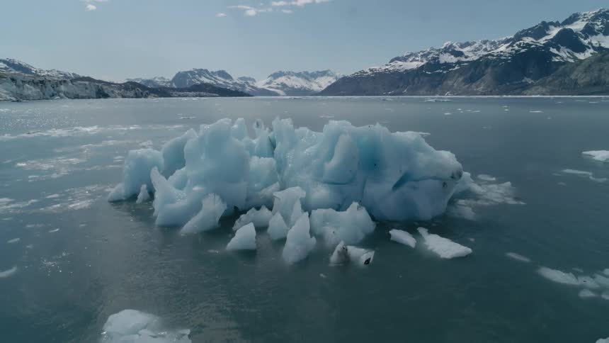 A large iceberg floats in a calm bay with snowy mountains around