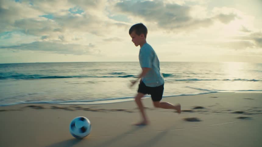 Boy running on the beach sand kicking a soccer ball