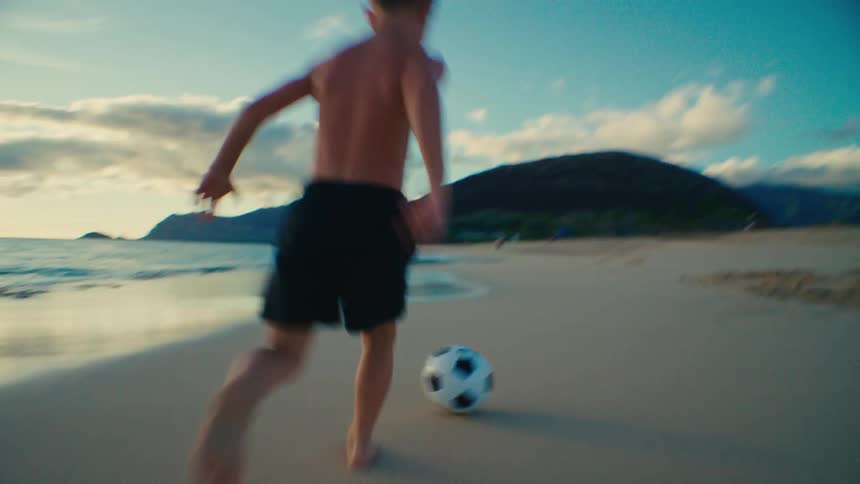 A kid in shorts continuously kicks a soccer ball around the beach sand at midday as the waves hit the shore