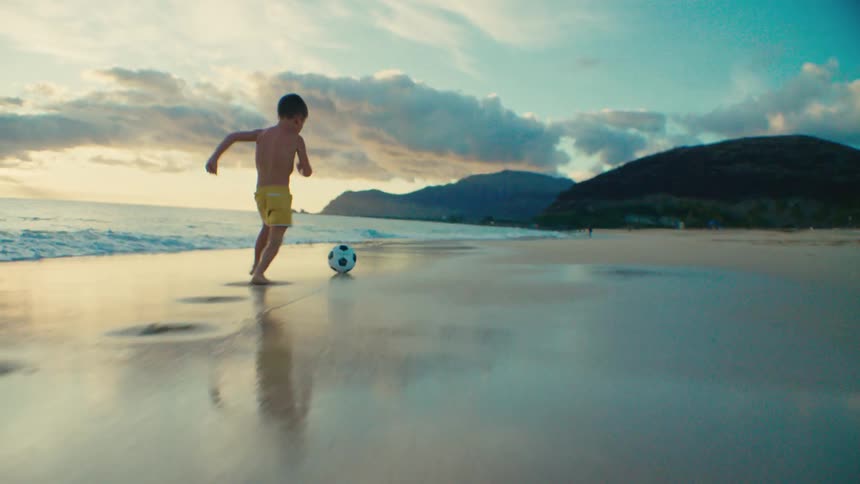 Young boy runs while playing a soccer ball on the beach at sunset