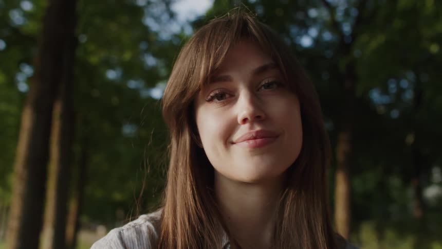 A woman with long hair stands alone in a sunlit park clearing surrounded by trees and soft shadows